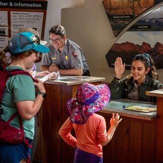 A park ranger sitting at a desk smiles while faces a young visitor. Both are raising their right hand.