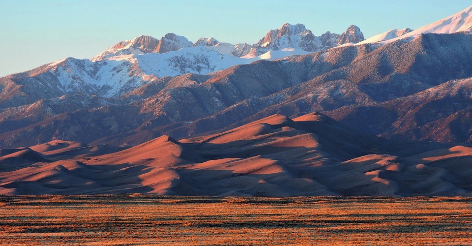 Snowy mountain peaks rise above a giant field of sand dunes casting long shadows during sunset