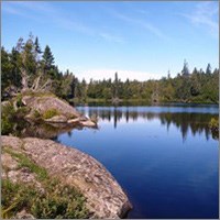 A blue lake reflects forest under a blue sky with rocky outcrops to left