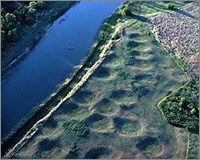 Aerial view looking down at river with green grassy field alongside. Field contains over a dozen large round depressions