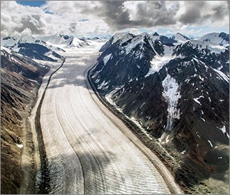 Aerial view of large valley glacier shining bright white with rocky, snowy peaks surrounding