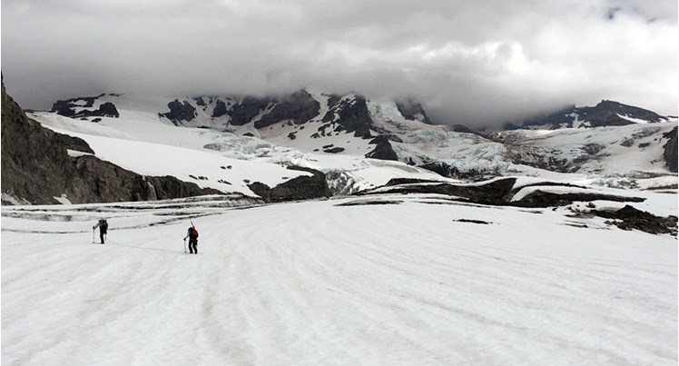 Two researchers walking on glacier with mountain in distance hidden behind clouds