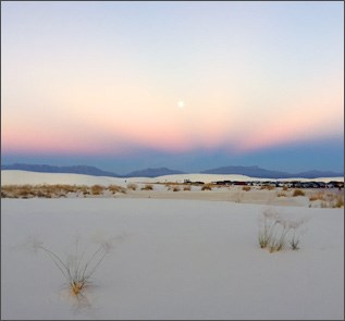 A setting sun is visible in a sky of purple, orange, and blue over dark mountains and a white field of dunes