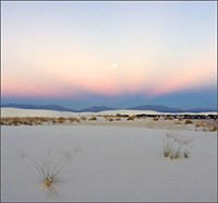 A setting sun is visible in a sky of purple, orange, and blue over dark mountains and a white field of dunes