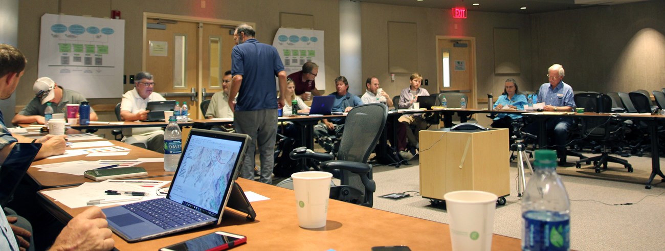 A dozen people sitting around conference room tables arranged circle