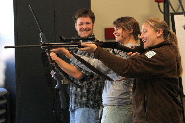 Three people stand shoulder to shoulder; two have guns raised up in a shooting position, and the third holds his gun pointing upward while smiling at the other two.