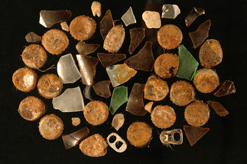 A sample of trash taken from the crop of a dead condor chick. Rusted bottle caps, aluminum can pull tabs, and broken glass laid out flat on a black background.
