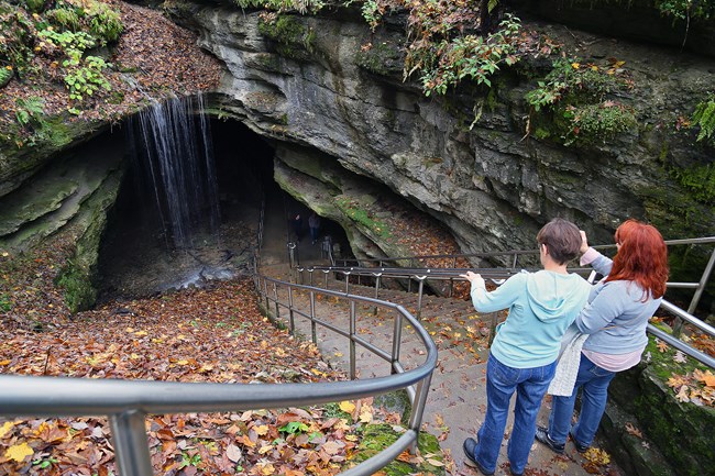 two people stand at top of stairs looking down at waterfall over the entrance to a cave
