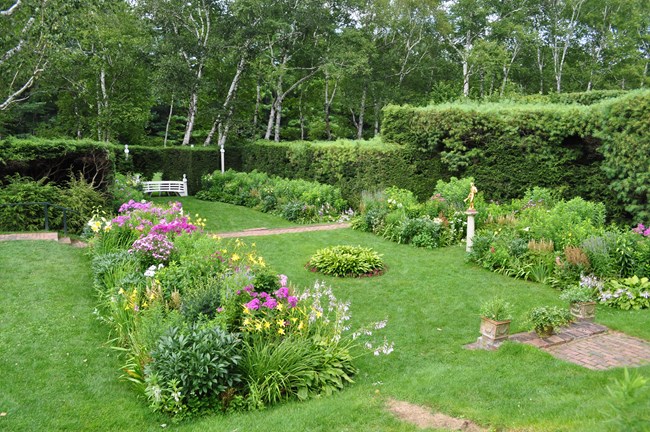 Garden at Aspet Short grass grows between rectangular planting beds with blooming perennial flowers, with brick walks and sculptures. It is surrounded by a tall hedge.