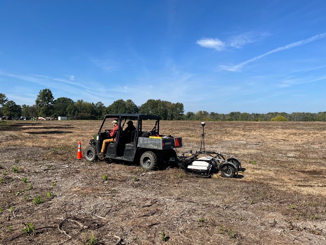 MAVA-GPR-Crew-Scanning-Site- Two people operate ground scanning equipment in an open field, driving a cart with a radar device attached to a trailer on the back.