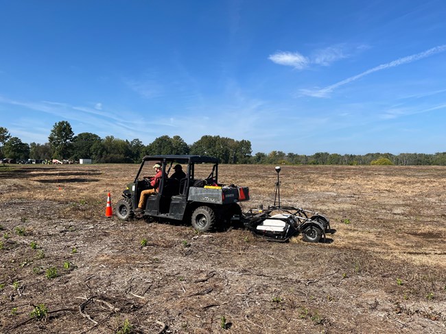 Two people operate ground scanning equipment in an open field, driving a cart with a radar device attached to a trailer on the back.