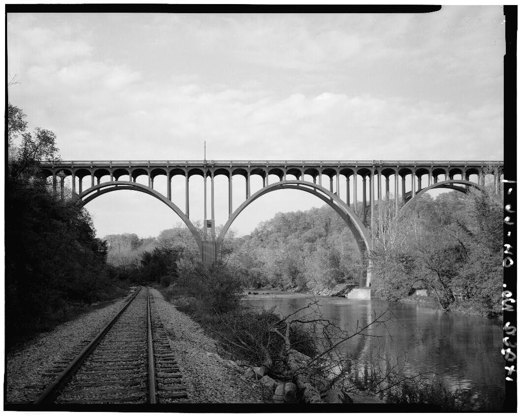 Tall, multi-arched bridge spans hillsides, crossing a railroad track and a river