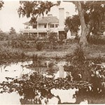 150_150-CUIS Water plants grow in a small pond with a two-story cottage beyond, historic structure