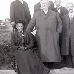 A woman in a long, dark dress seated beside standing men. A woman in a long, dark dress seated beside standing men.