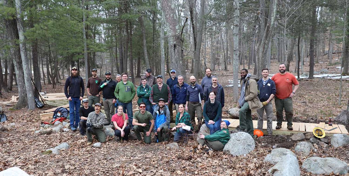 A group of people pose in front of boardwalk construction in the woods in early spring