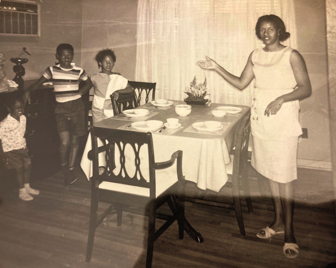 Myrlie Evers and children in dining room, around 1963. Myrlie Evers stands beside a table set with dishes and silverware, one arm outstretched. Three children stand on the opposide side of the table. Long, sheer curtains cover the window. (sepia-toned)