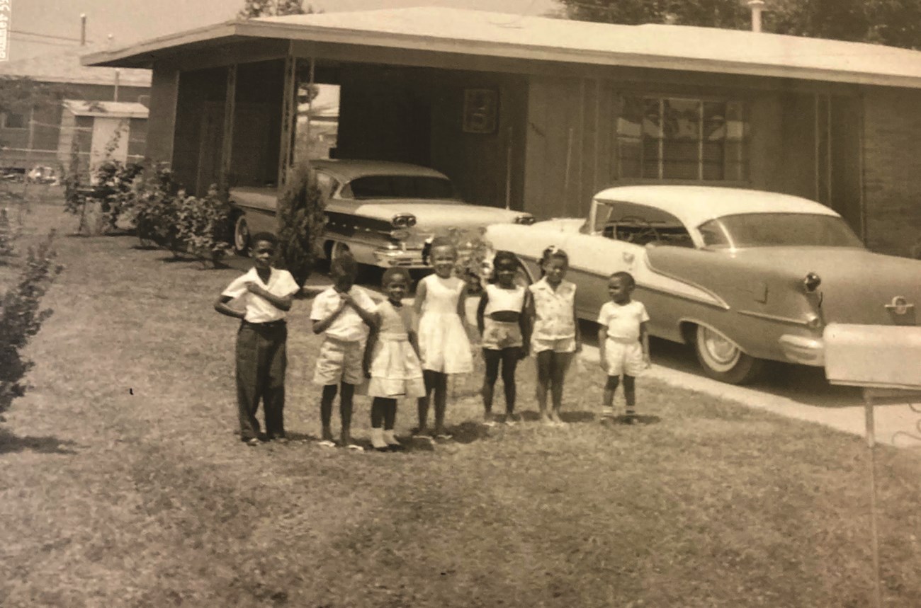 Evers children and neighbors in the yard, summer 1959 Seven African American children stand side-by-side in the grass beside two 1950s-era cars in the driveway of a ranch house, sepia-toned photo.