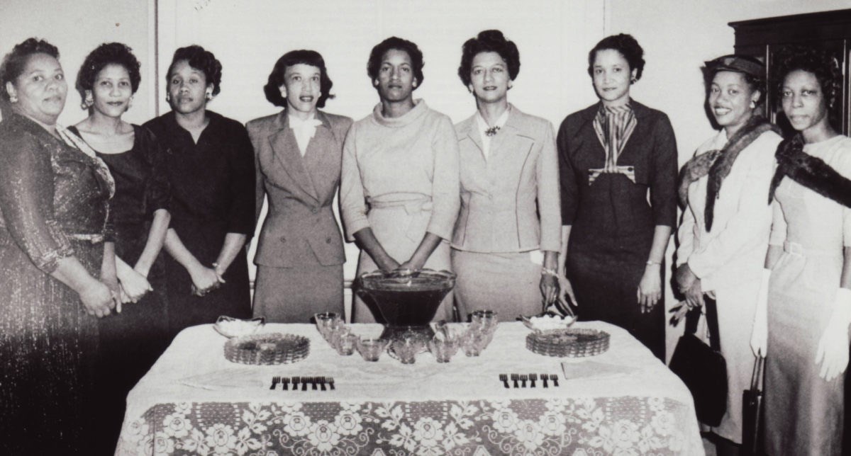 Nine African American women stand behind a table with a symmetrical layout of punch bowl, cups, forks, and plates.