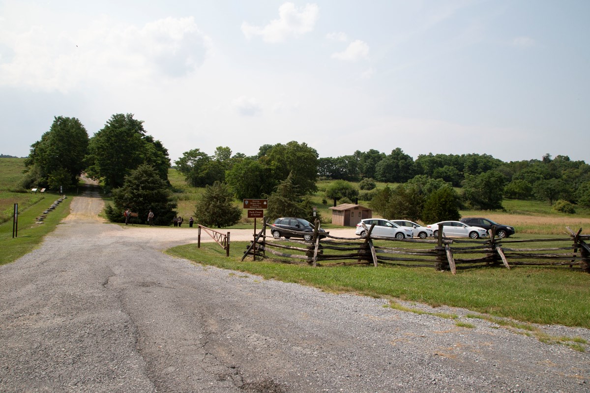 A gravel road leads through rolling terrain with scattered trees. A wooden fence, parking lot, and small comfort station are to the right of the road.