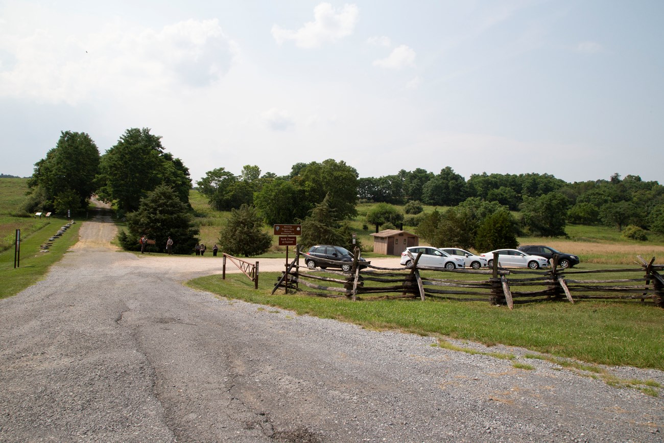 Road and parking lot at School House Ridge North A gravel road leads through rolling terrain with scattered trees. A wooden fence, parking lot, and small comfort station are to the right of the road.