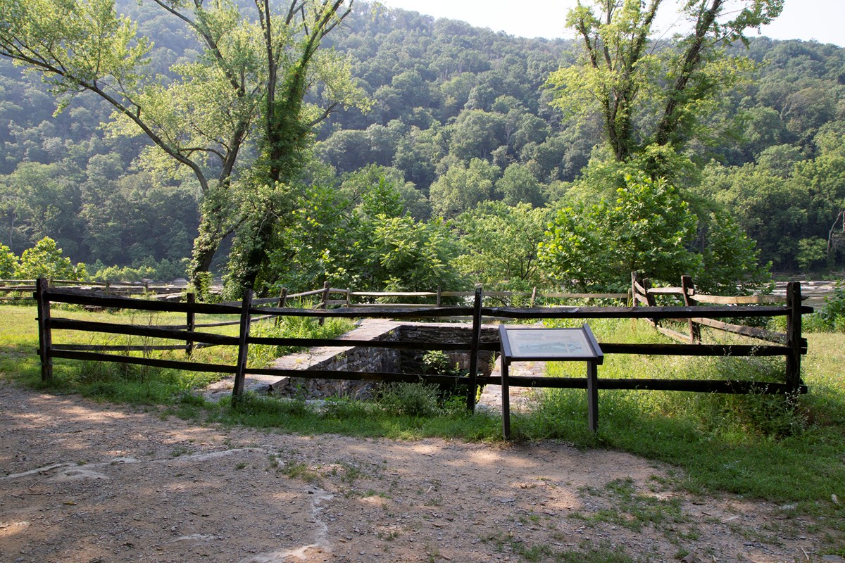 An interpretive sign in front of a rectangular, stone-lined tunnel, surrounded by a wooden fence.