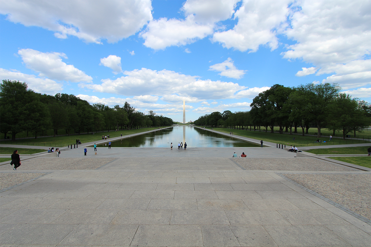 View from Lincoln Memorial landscape The vertical Washington Monument stands at the far end of a rectangular pool of water, reflecting clouds and surroundings. Trees and turf line the sides.