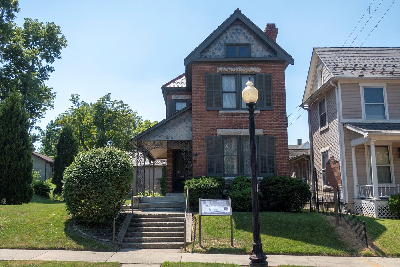 A neat yard of lawn and shrubs surrounds a brick house with a porch opening up to the left side