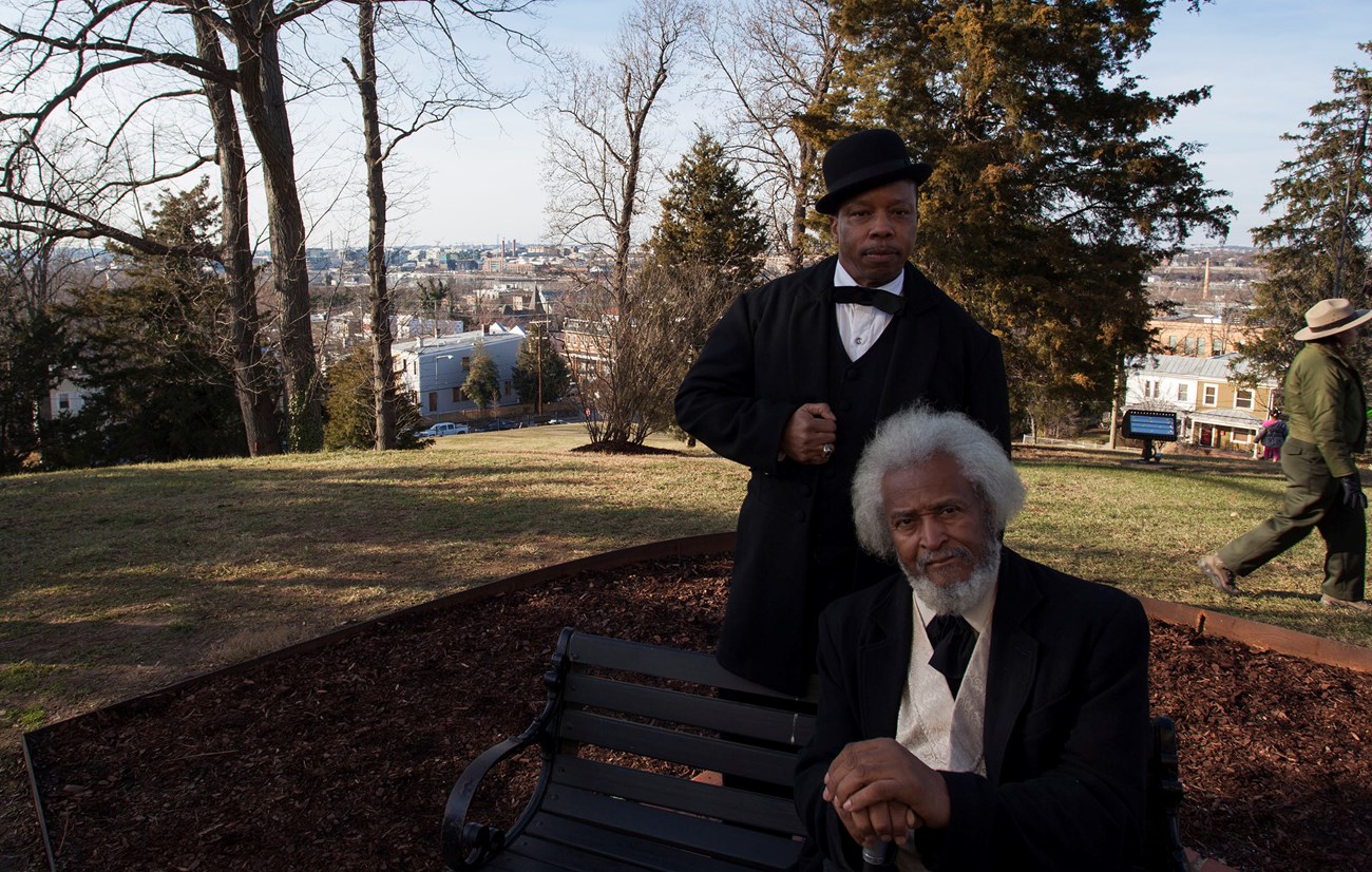 Two African American men in suits, one standing and one seated on a bench, atop a sunny hill with a view of a city beyond.