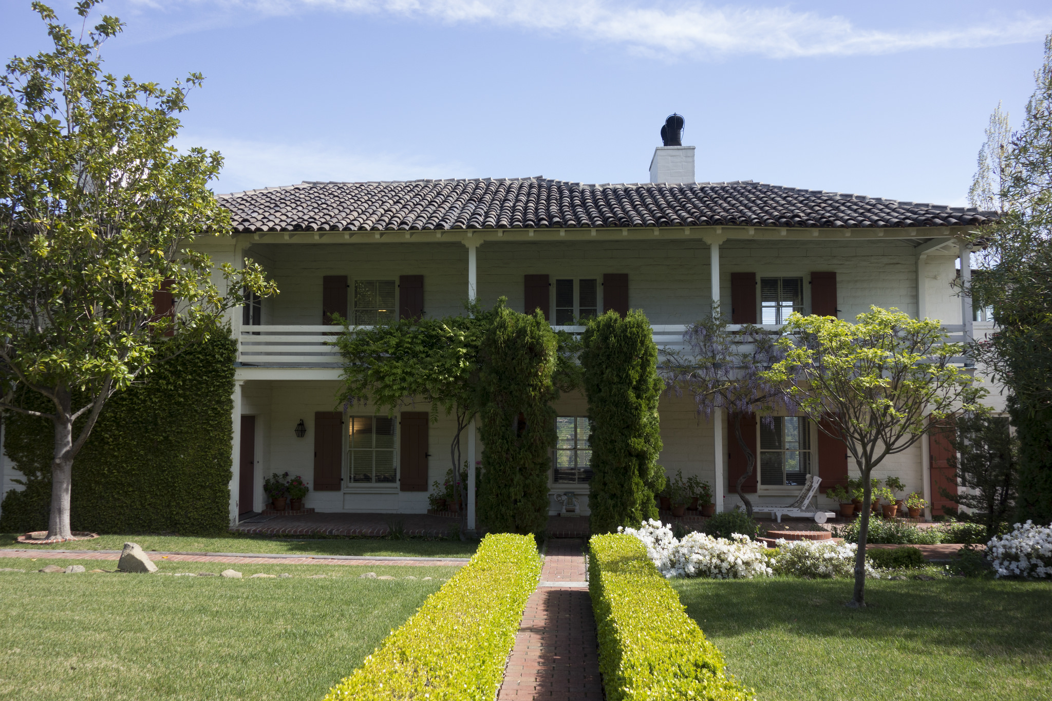 A straight path between boxwood hedges and manicured courtyard leads to a two-story house with porches on each story