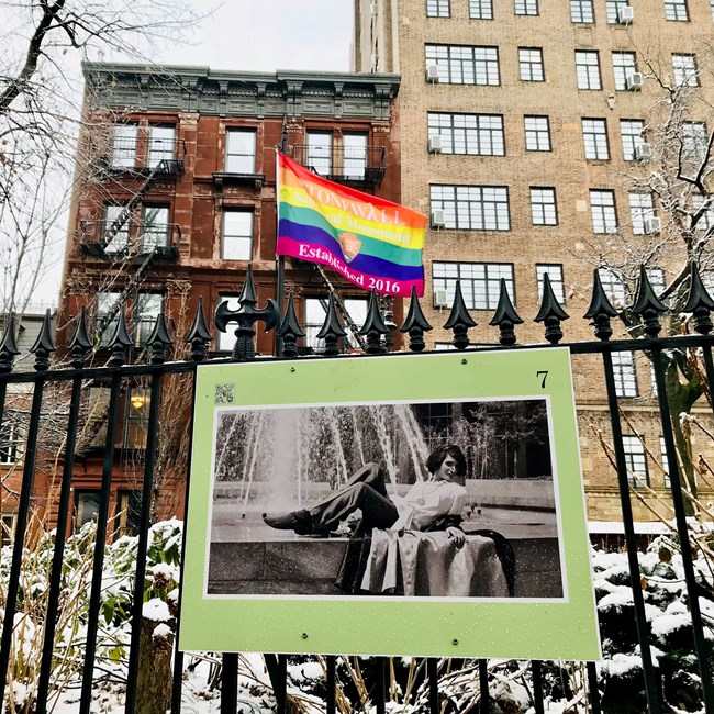 A photo of Silvia Rivera hangs on a metal fence in a snowy park, with brick buildings and a rainbow flag in the background.