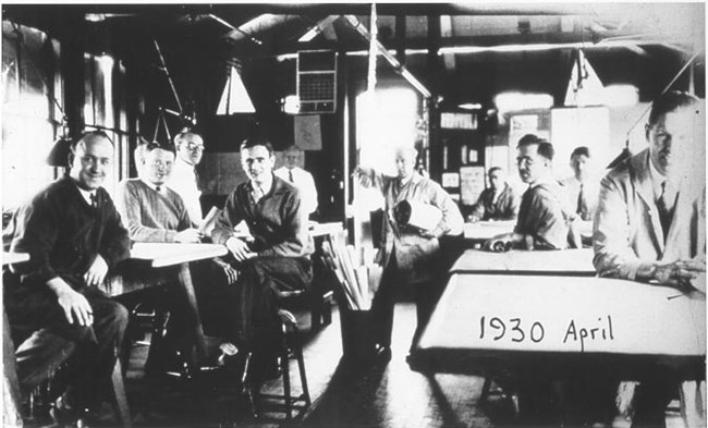 A group of men are seated around tables. A container with rolled up papers is on the floor between them. Photo is labeled April 1930.