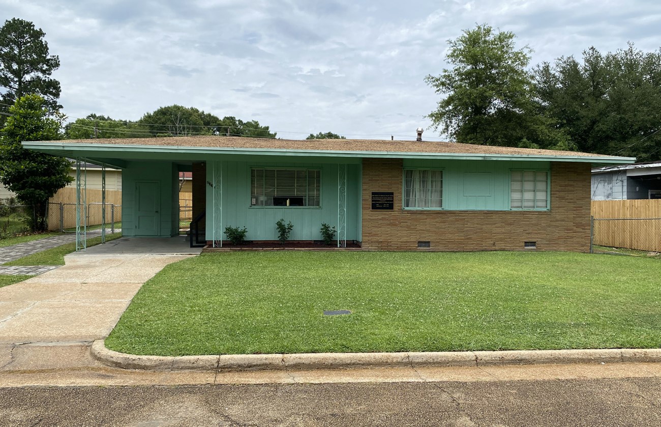 Front of Medgar and Myrlie Evers House NHL from street A flat, open lawn connects the road and curb to a one-story ranch house with driveway and carport to the left.