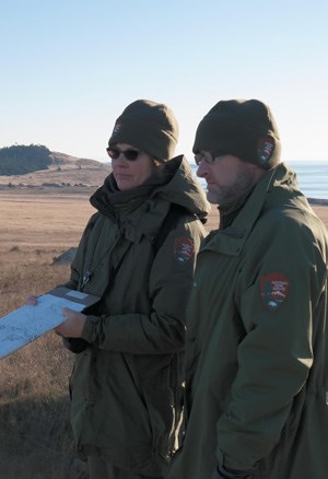 lt= Two NPS cultural resource professionals are dressed for cold weather on a sunny day in an open, grassy landscape.