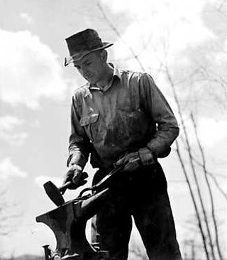A low angle of a blacksmith in a hat and button down shirt, looking down at a hammer and anvil.