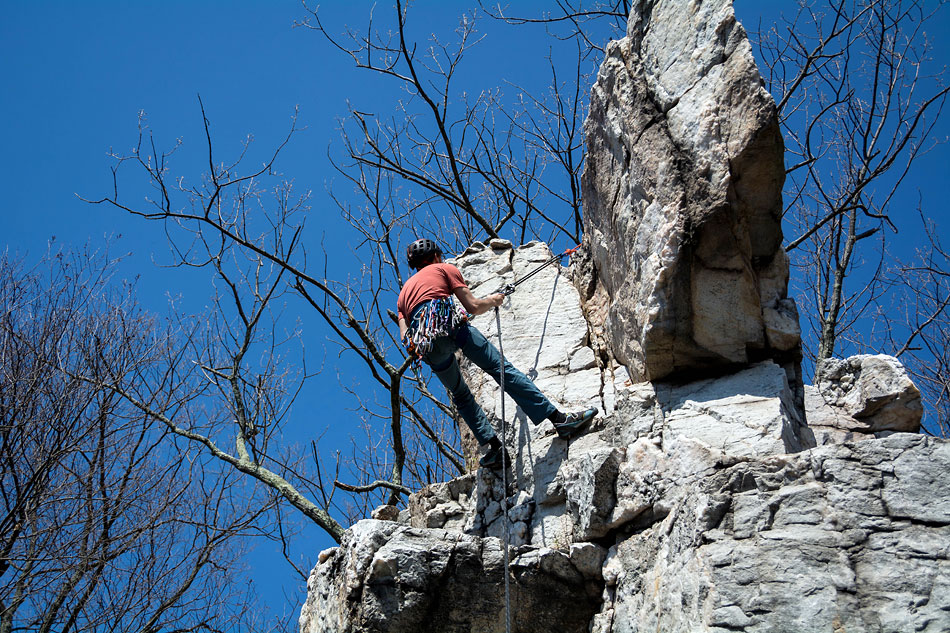 A climber, wearing a helmet and with gear hanging from his harness, hangs from a rope attached to the top of a rocky prominence.