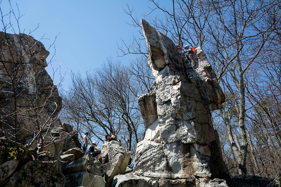 A group of climbers with ropes and gear around the base and top of jutting rock, under leafless trees and blue sky.