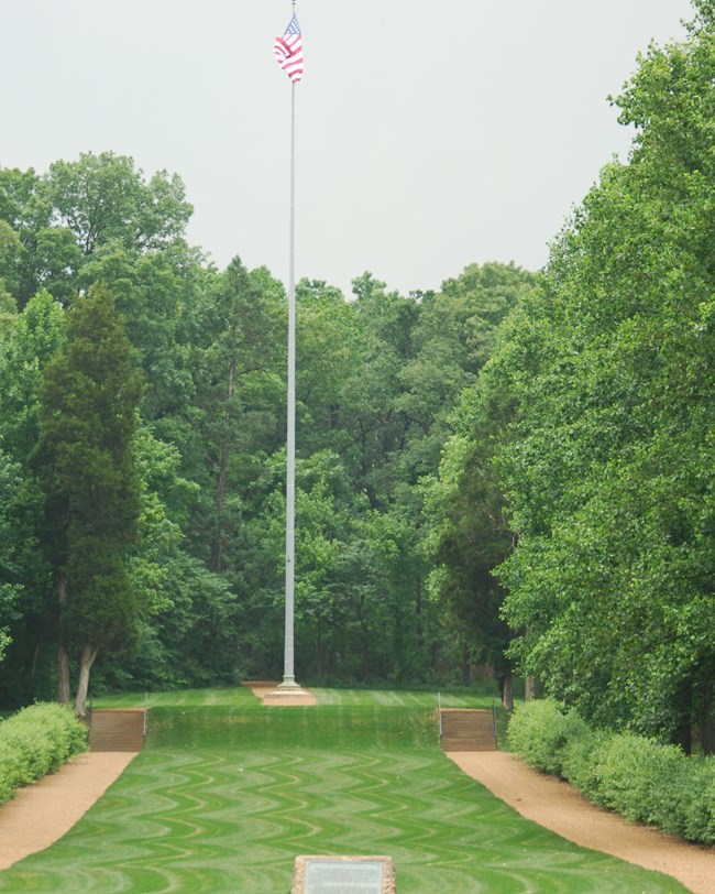 400-500-LIBO-allee Leafy trees line the sides of an areas of neatly mowed turf, with a tall flagpole at the end