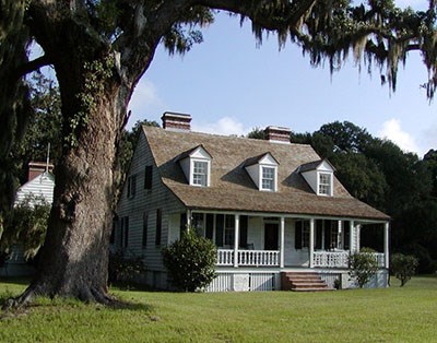 A tall tree with Spanish moss towers over a farmhouse, with a front porch and three dormer windows.