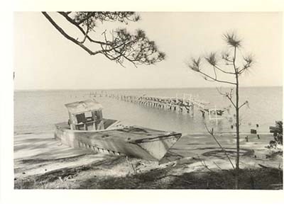 The ruins of a wooden steamboat rest on a sandy beach, with a wooden wharf extending into the water.