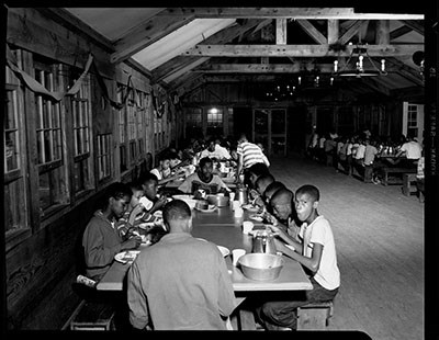 Historic image of the dining hall interior at Cabin Camp 4 shows African American boys seated around tables at meal time.