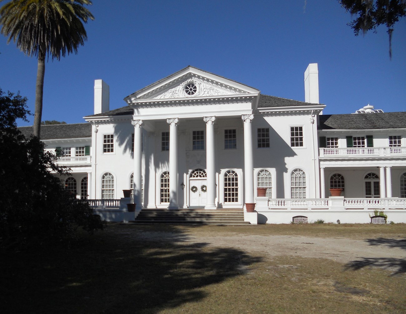 A two-story mansion has four columns across the front, a curving entrance road, chimneys, and rows of windows.