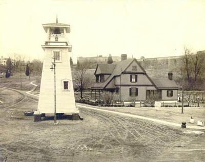 A squat, rectangular lighthouse stands near a large house, with fort walls on the hill beyond
