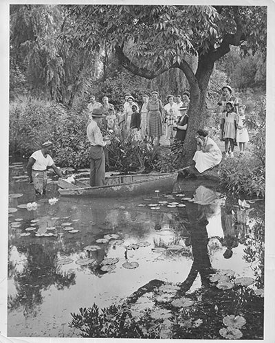 A park ranger stands in a rowboat near the edge of a pond as he addresses a small crowd.