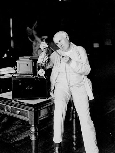 Edison and his Home Projecting Kinetoscope A man, seated on the edge of a table, furrows his brow as he looks at a strip of film that he holds up between two hands.