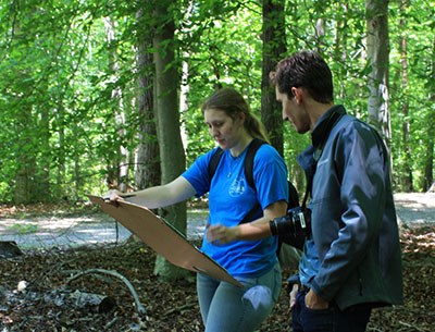 One student with a camera around his neck looks over the shoulder of a student as she draws on a board, under a canopy of trees.