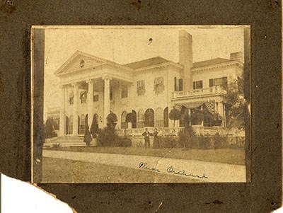 Three people stand on the lawn in front of a grand two-story house with four columns at the front and striped bunting over the windows.