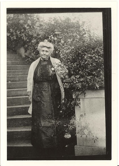 A woman stands at the base of a staircase beside flowering vines, wearing a long dress and her hair in a loose bun.
