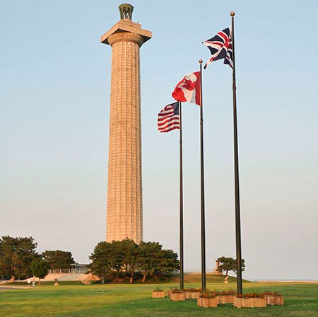 450-PEVI_memorial Three flagpoles in a line in the grass in front of a memorial column with US, Canadian, and British flags