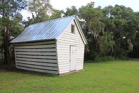 Wooden structure with one door and a peaked roof at the edge of a lawn, with moss-draped trees beyond.