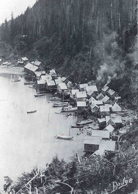 Smoke rises from the chimneys of a tight cluster of structures along a steep, wooded shoreline with boats in the water - black and white